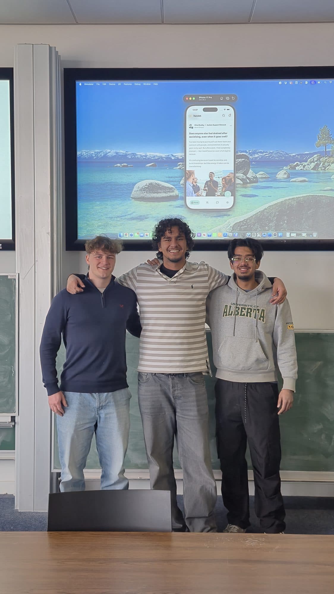 Three team members standing in front of a large screen showing the application running in an iOS simulator.